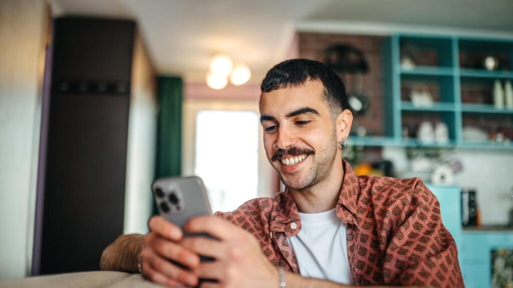 Cheerful hipster man using smart phone while relaxing on a sofa at home