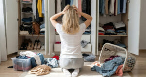 Stressed woman looking at messy closet full of clothes with laundry baskets on the floor at home